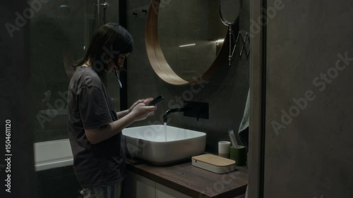 Medium full side shot with VFX effects of anxious young girl standing in bathroom, brushing teeth while scrolling social media, receiving malicious messages and threats from online stalker