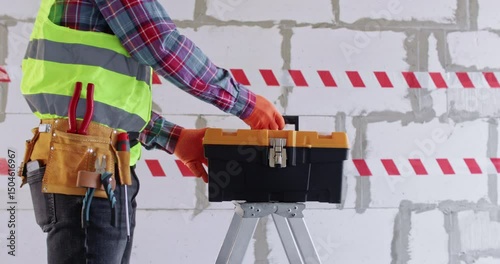 Engineer places toolbox on stepladder in large apartment hall. Male builder walks against bright warning tape and brick wall starting to work at renovation