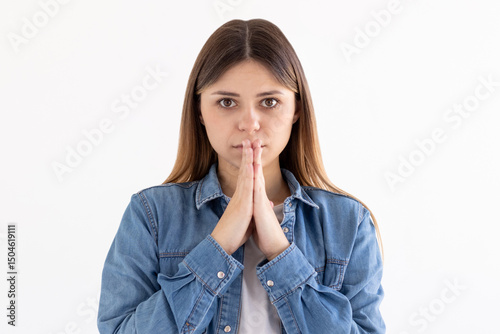 Portrait of imploring girl tilts head, purses lips, looks sadly at camera, makes praying gesture, asks for help and support, begs boyfriend for apologize, wears jeans shirt, isolated on white wall