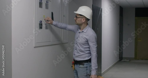 Foreman in helmet inspects electrical panel to check wiring connections. Specialist in glasses evaluates special devices condition before carrying out flat renovation