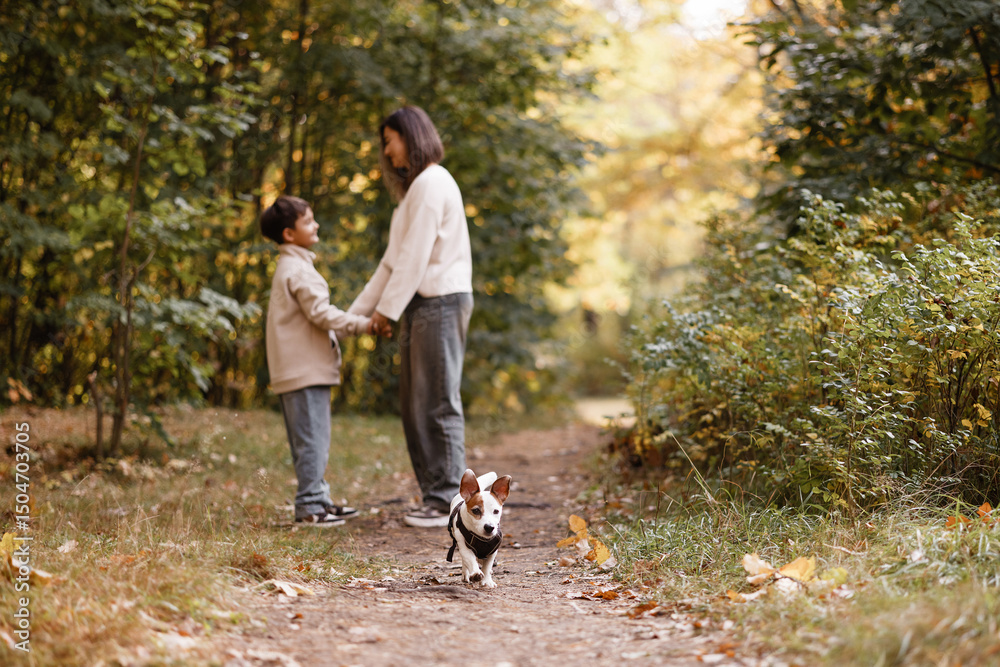 Fototapeta premium Side view of an Asian mother and her young son laughing while walking a small white dog on a forest trail. They wear casual autumn clothes. Great for family, parenting, and outdoor lifestyle themes.