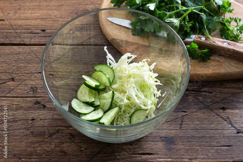 Fresh cucumber and cabbage salad in glass bowl on wooden table, healthy vegetarian food preparation concept
