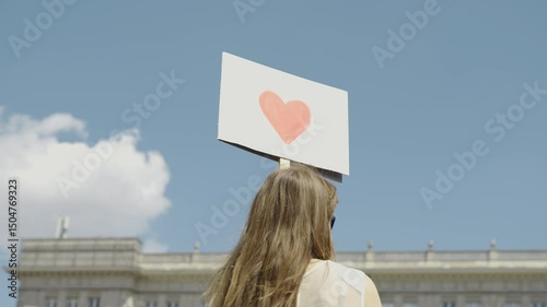 Young woman standing in an urban environment holding a sign with a red heart during a peaceful demonstration. A symbol of hope, unity, and emotional connection in a public space — ideal for social awa
