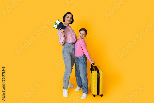 Family Travel. Happy Young Mother And Daughter With Tickets And Suitcase Posing Over Yellow Studio Background, Cheerful Mom And Female Child Ready For Vacation Trip, Full Length, Copy Space