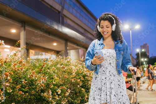 Young Latin woman traveler smiling near a bus terminal, listening to music with wireless earbuds, holding backpack and checking her phone