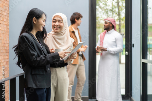 Muslim businesswoman in hijab discussing with asian colleague working on tablet in office, teamwork in a modern corporate environment, successful women collaborating on business projects, technology