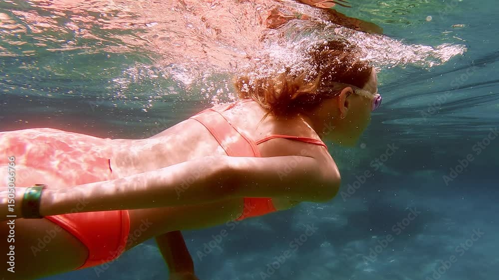 Woman swimming gracefully underwater in clear blue sea, generating bubbles and luminous reflections while sunlight radiates across her moving body