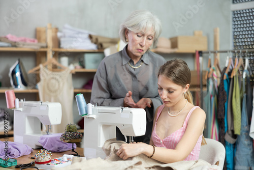 Mature woman expressing dissatisfaction about work of young tailoress sitting at sewing-machine in tailor's workshop photo