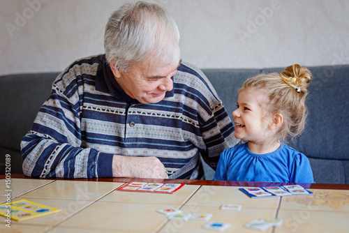 Beautiful toddler girl and grandfather playing together pictures memory table cards game at home. Cute child and senior man having fun together. Happy family indoors
