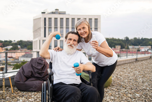 Senior couple practicing fitness on terrace. Elderly man with dumbbells supported by a woman holding water, promoting health, companionship active lifestyle and rehabilitation for mature individuals.