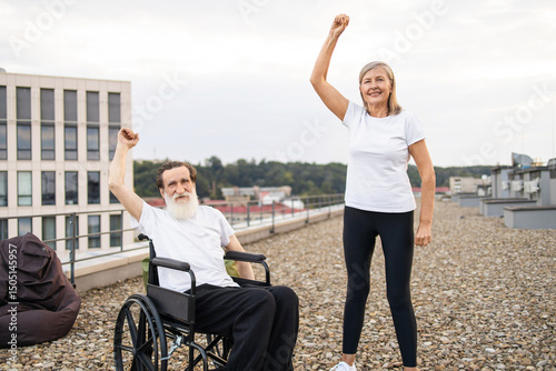Elderly man in wheelchair and woman standing together lifting arms outside urban rooftop