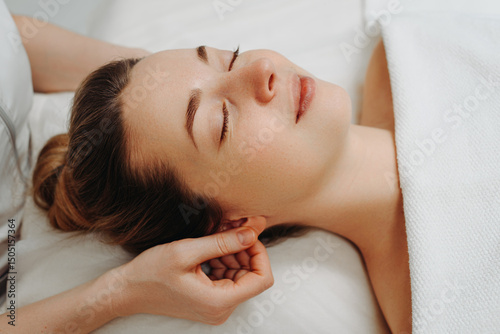 face massage, young attractive caucasian woman, beautiful relaxed lady, white towel, spa therapist doing facial and head massage in beauty salon, massaging ears, skincare and bodycare concept