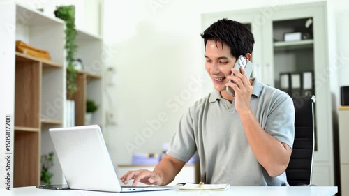 Confident Asian man talking on smartphone and working on laptop at office desk.