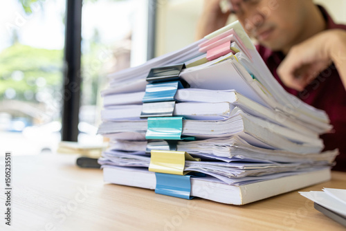 A man in a red shirt is sitting at a desk with a pile of papers in front of him. He is stressed and overwhelmed by the amount of work he has to do
