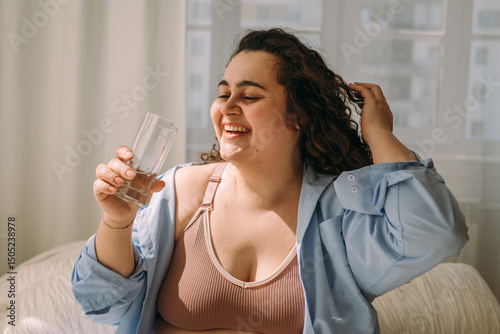 A positive young curly-haired plus-size woman drinks water after waking up in bed.Healthy lifestyle.