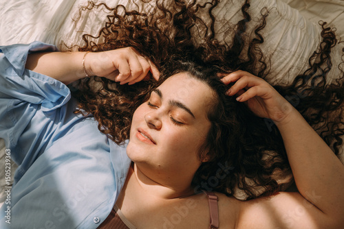 A portrait of a young, attractive plus-size woman with dark curly hair lying in bed with her eyes closed.
