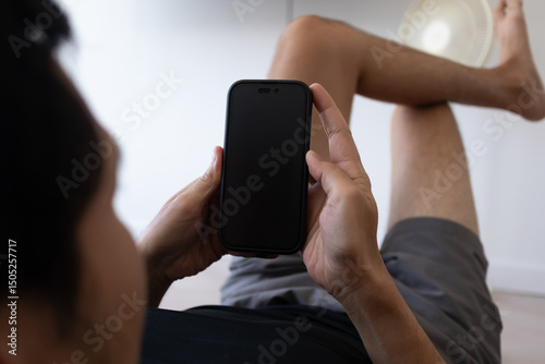 Asian man holding smartphone showing empty black sceen while lying on floor indoor.