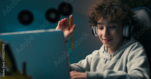 A boy with curly hair sits at a desk wearing wireless headphones, taking notes and redrawing. He looks off to the side, wondering about the next step in his school task.