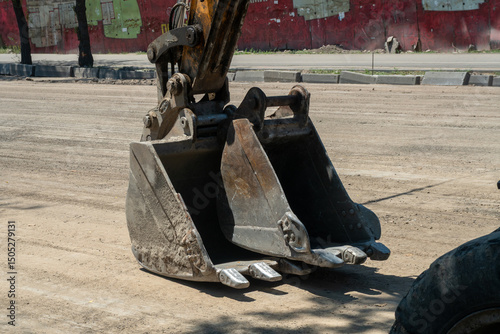 Heavy excavator bucket positioned on dusty street, engaged in grading, digging, and material removal as part of roadbed preparation for upcoming asphalt paving in city environment