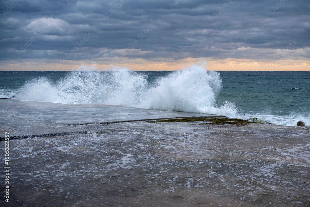 Obraz premium Stormy sea waves crashing against a rocky shore at sunset. Dark clouds fill the sky, creating a dramatic atmosphere with vibrant colors on the horizon.