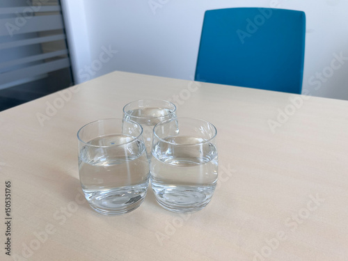 Three clear glasses filled with water sit on a light wooden table in a minimal office setting.