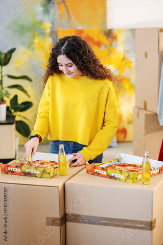 Young woman in yellow sweatshirt cuts pizza on moving boxes. She takes a break during relocation, enjoying a meal amidst cardboard boxes and belongings.