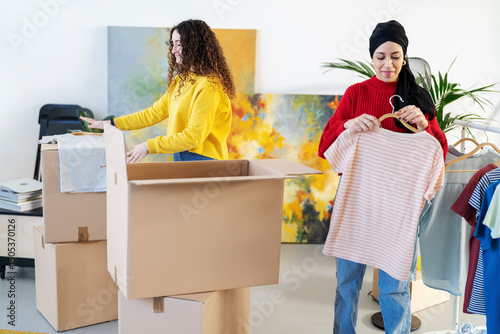Two women are packing clothes into boxes for moving or storage. One woman is placing items into a large box, and the other is holding a shirt on a hanger.