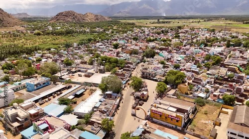 A bright aerial view of an Indian town nestled beside lush green fields and rocky hills, with colorful houses, busy streets, and distant mountain ranges.