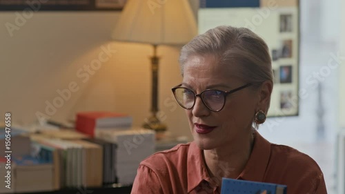 Medium closeup of mature Caucasian female author in glasses speaking thoughtfully during bookstore event, surrounded by stacks of books and warm lighting in intimate literary setting