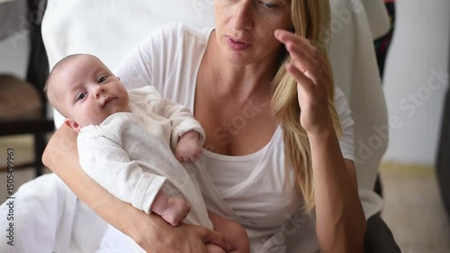 Blonde mother in white T-shirt sits on armchair and holds smiling baby in white overalls on hands. Newborn - happiness for family. New worries and lifestyle.