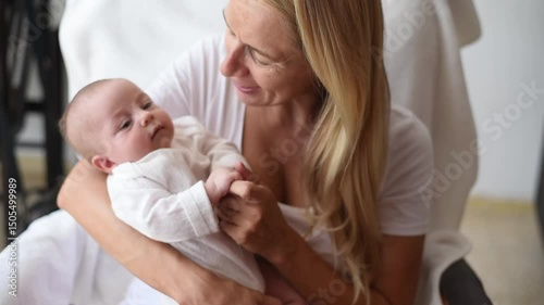 Mother-blonde in white T-shirt sits on armchair and holds smiling baby in white overalls on hands. Newborn - happiness for family. New worries and lifestyle.