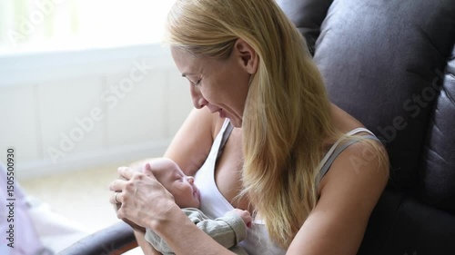Mother-blonde in white T-shirt sits on armchair and holds sleeper baby in olive overalls on hands. Newborn - happiness for family. New worries and lifestyle.