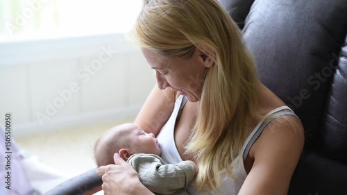 Mother-blonde in white T-shirt sits on armchair and holds sleeper baby in olive overalls on hands. Newborn - happiness for family. New worries and lifestyle.