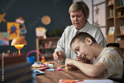 Young boy with disability engaging in creative activity with help from teacher in classroom setting. Teacher supervising and assisting student with crafts at wooden table