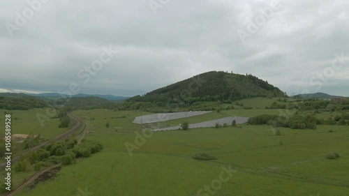 Wide aerial shot of solar panels in neat rows on green terrain at the foot of forested Chełmiec mountain in Boguszów-Gorce under a gray overcast sky