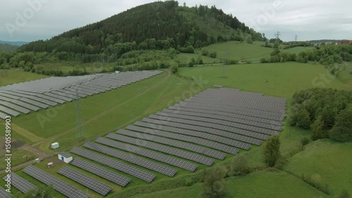 Aerial drone view of solar panels arranged in rows on open grassland with Chełmiec mountain and forest trees surrounding the site in Boguszów-Gorce