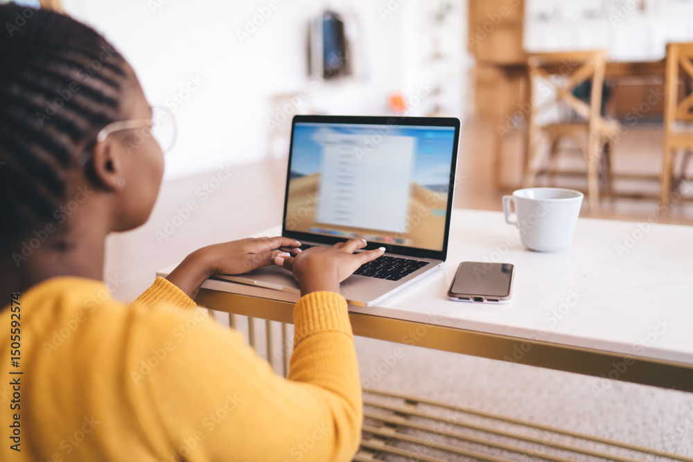 Young African woman typing on laptop with focused expression, working remotely at coffee table in cozy living room with smartphone and cup nearby, immersed in digital task Young African woman typing on laptop with focused expression, working remotely at coffee table in cozy living room with smartphone and cup nearby, immersed in digital task