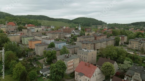Drone shot of a quiet residential neighborhood in Boguszów-Gorce, Poland featuring rooftops, trees, and a peaceful road surrounded by green hills and nature