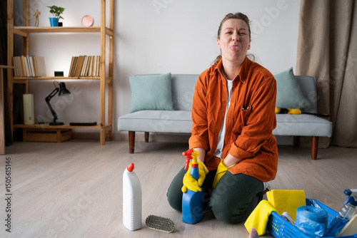 Positive woman wearing yellow gloves sitting on floor with cleaning supplies and detergents, showing tongue-out. Home cleaning, household, housekeeping and housework