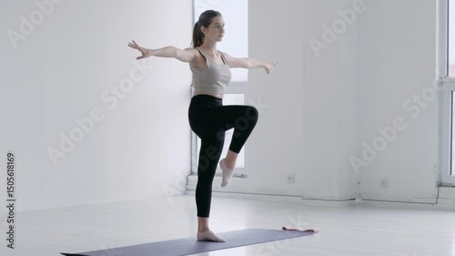 Gorgeous caucasian young female in sportswear doing warm-up before yoga In a bright studio with windows. Girl stands on one leg and rotates the other. Balance and equilibrium of the body exercise