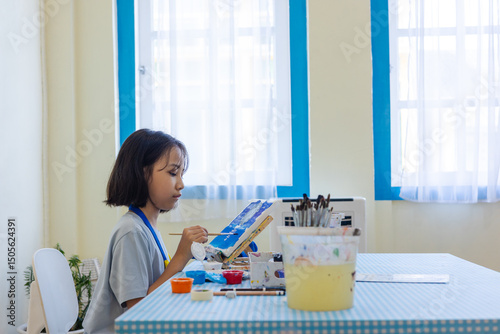 Young Asian student deeply in a painting workshop, brushing colors onto a canvas. Indoor pastel-toned classroom enhances the calm, focused mood. Surrounded by tools and paints for hands-on learning.