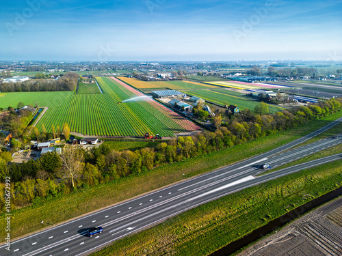 Aerial panoramic view of the Dutch countryside featuring farmland, green fields, forests, and a highway stretching through the landscape toward a distant town.