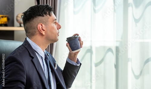 A man in a suit is drinking coffee and looking out the window thoughtfully.