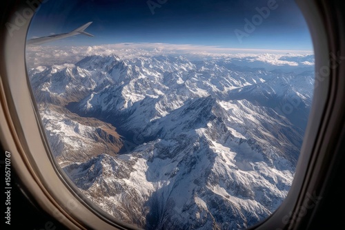 Looking out an airplane window at a dramatic mountain range below, with snow-covered peaks and a clear blue sky above the horizon.