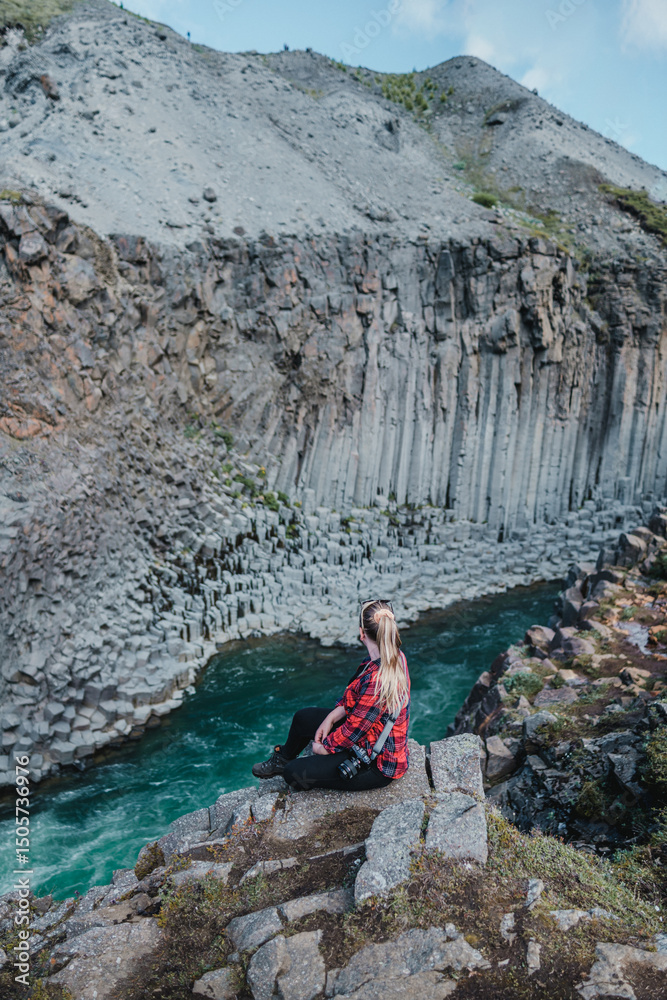 Naklejka premium Woman sits on cliff edge overlooking basalt columns in Studlagil Canyon...