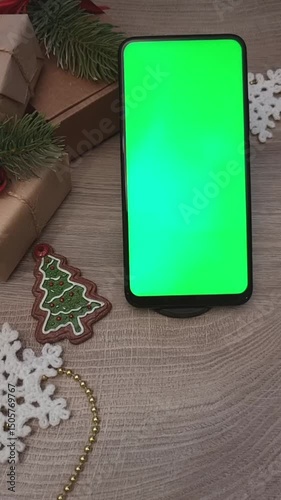 A smartphone with a green screen lies vertically on a wooden table next to gifts and homemade Christmas decorations. Vertical video.
