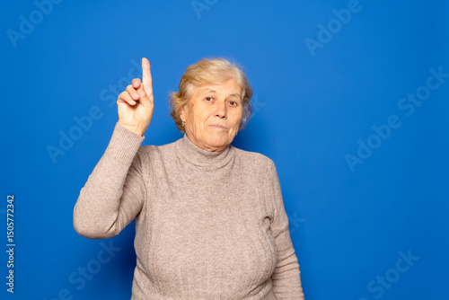 Photo portrait of a happy elderly Caucasian woman in her 70s having an idea and raising her finger, isolated over a pastel blue background