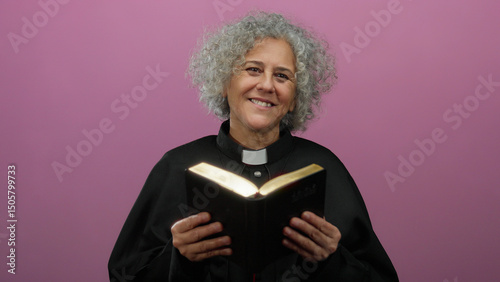 Fotografie Woman priest with grey hair reading a bible against a pink background
