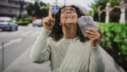 Middle-aged woman holding indonesian rupiah banknotes while smiling and pointing upwards in a busy city street. photo