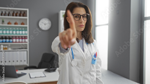 Wallpaper Mural Woman doctor gesturing one finger in a clinic room with shelves and clock in the background wearing glasses and stethoscope suggesting attention or warning alert indoors. Torontodigital.ca